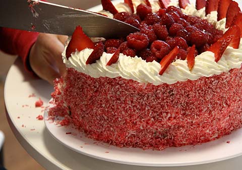 A red cake with fruit on top being cut with a knife